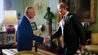 king charles iii shaking hands with chris hadfield in a palace room surrounded by photos, chairs, tables. a statue is on a pillar between them
