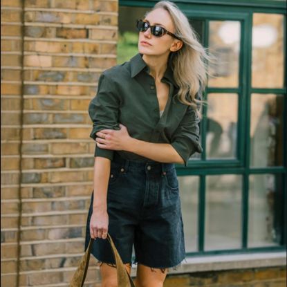 Middle-aged woman wearing black denim shorts, tailored green shirt, and sunglasses with wall in background.