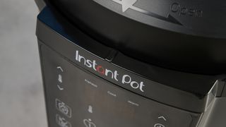 a black pressure cooker photographed against a blue background, showing its cooking presets, steam release valve, and Instant Pot branding