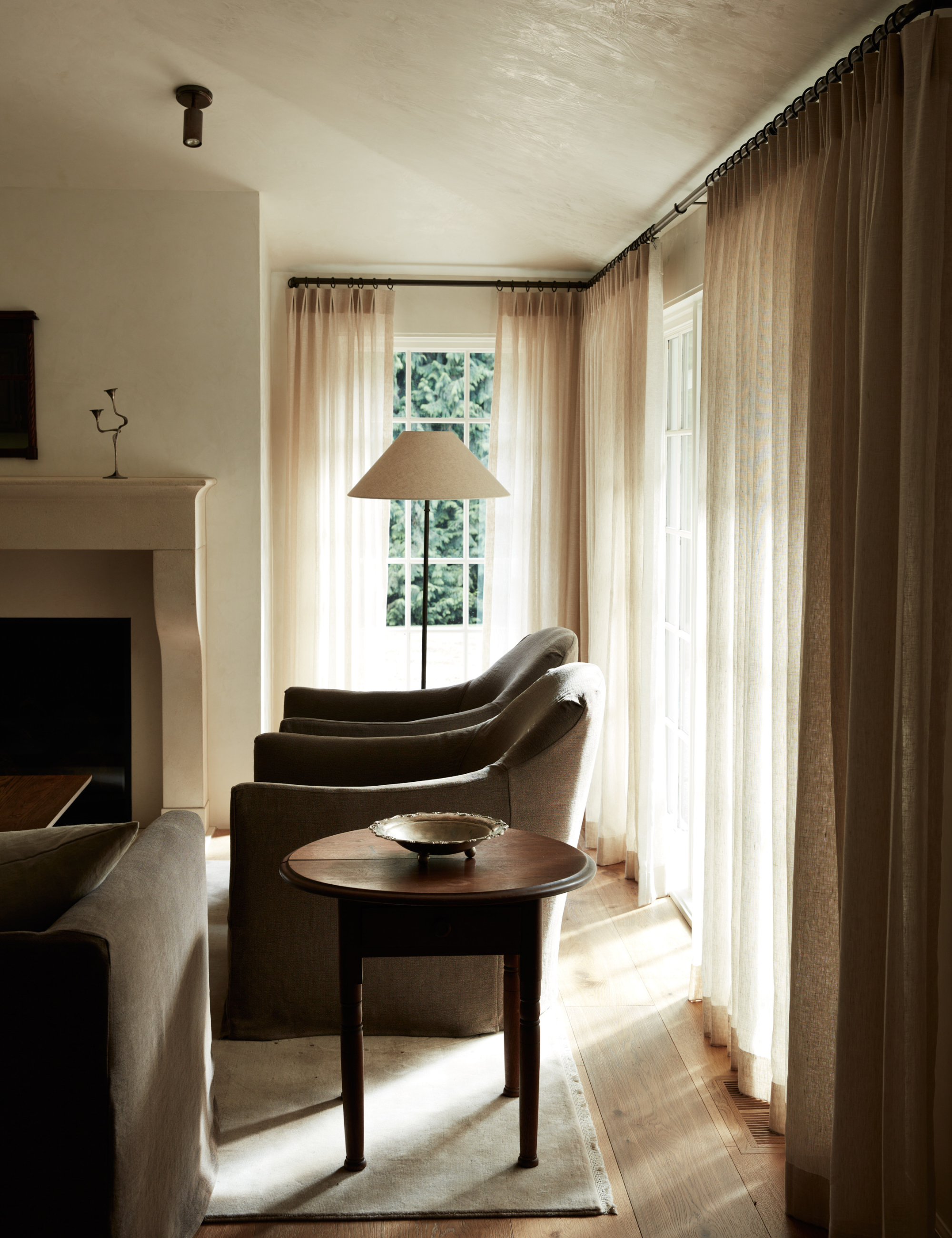 Sunlit warm minimalist living room featuring sheer curtain drapes, a linen-shaded iron floor lamp, olive green accent chairs, and a vintage wooden side table