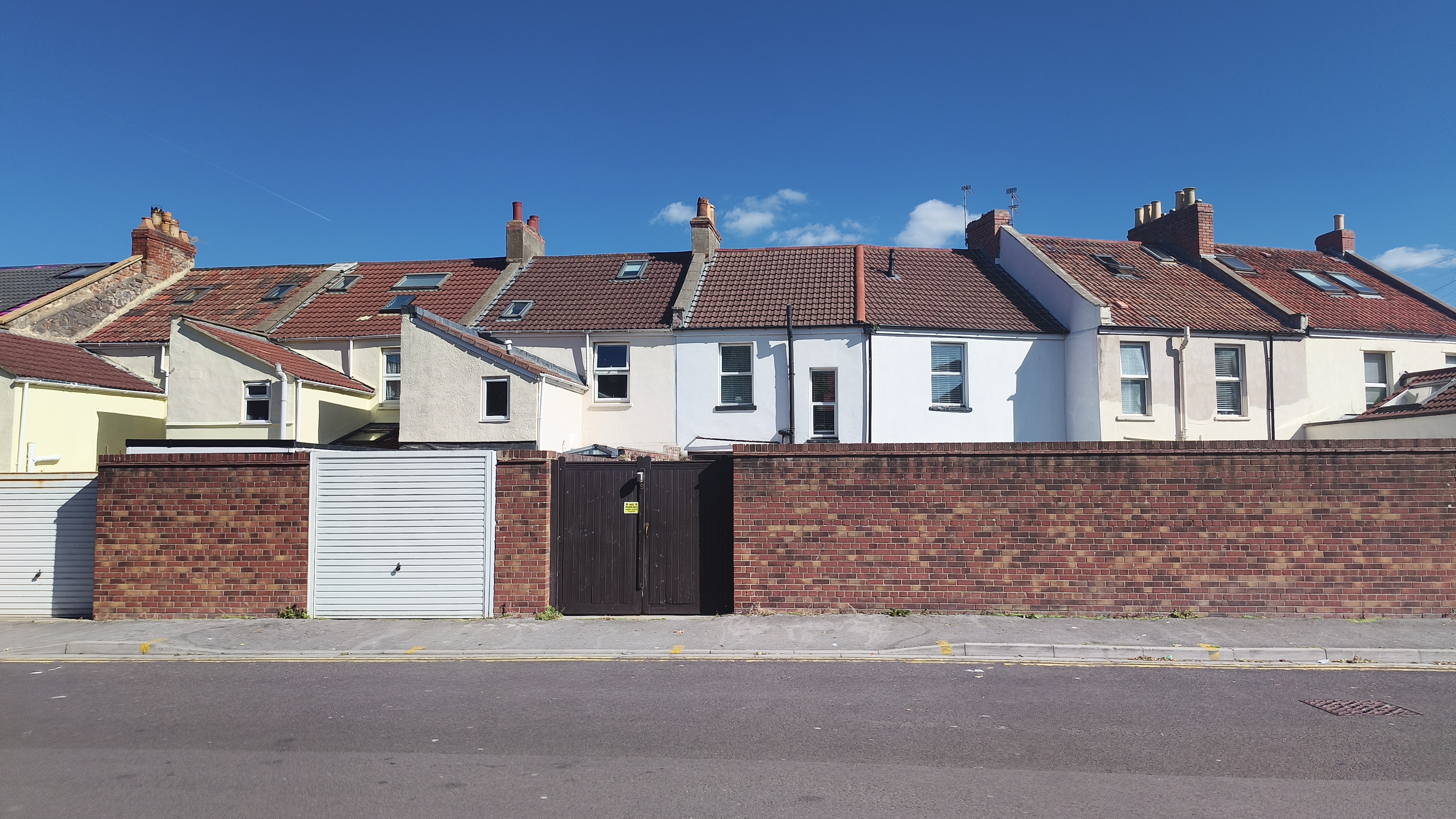A row of residential buildings with brick and white facades is seen from across the street, with a tall brick wall and garages in the foreground.