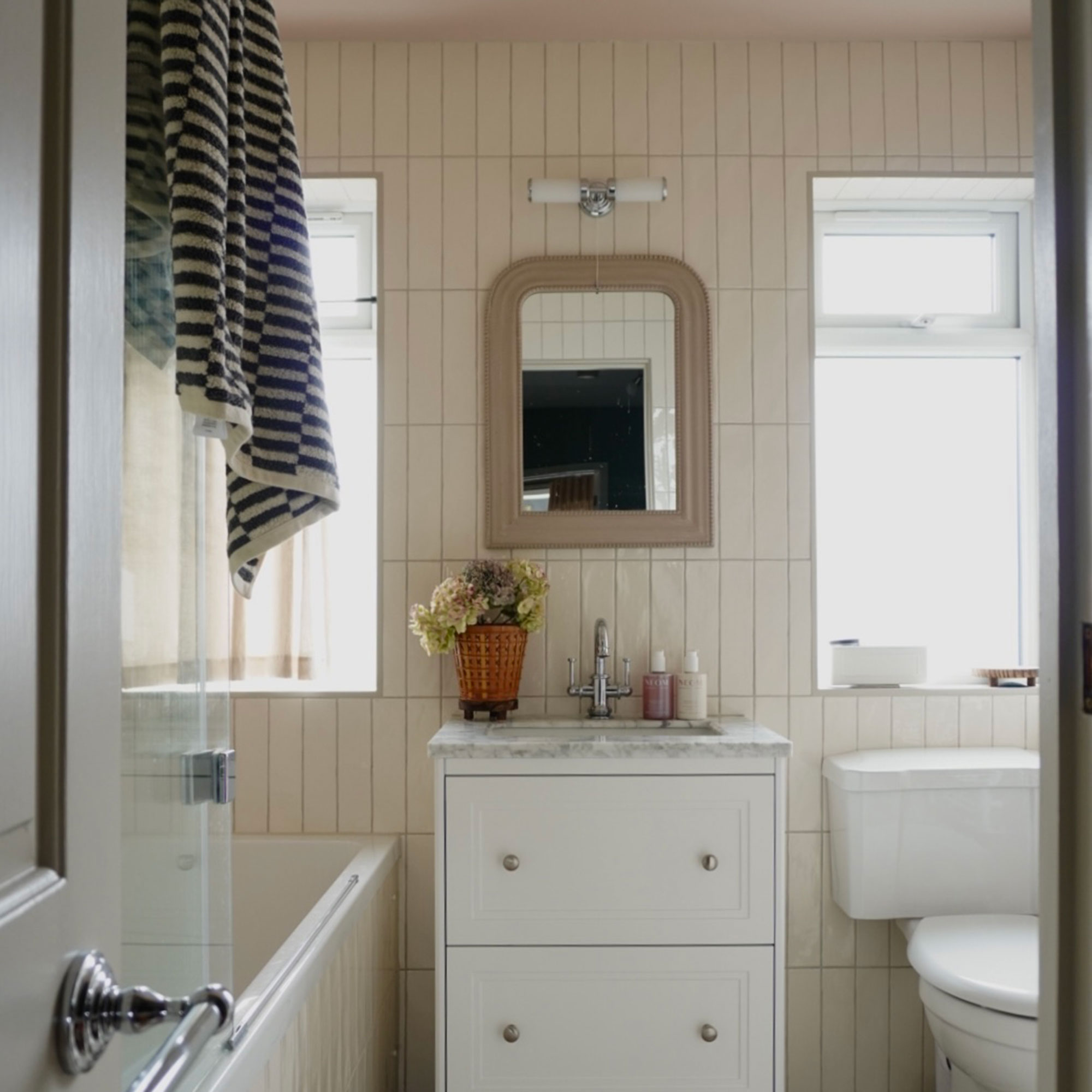 Neutral tiled bathroom with toilet, vanity unit, small mirror, bath and two windows