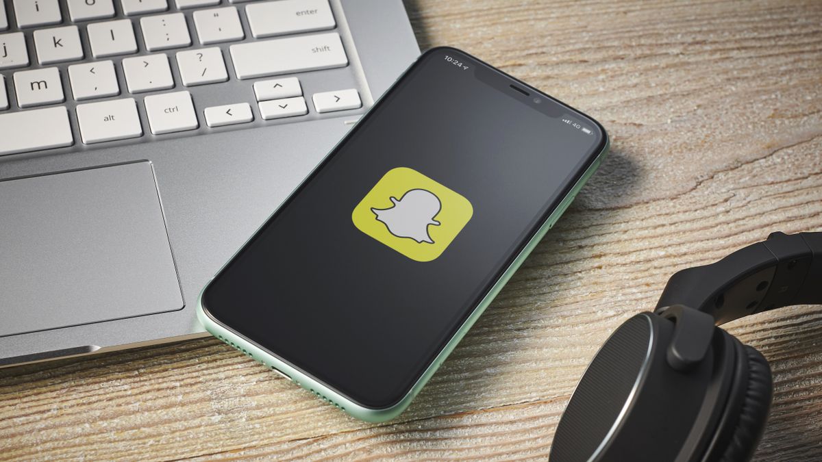 An iPhone, siting on a laptop keyboard next to a pair of headphones, with the Snapchat messaging app logo on screen