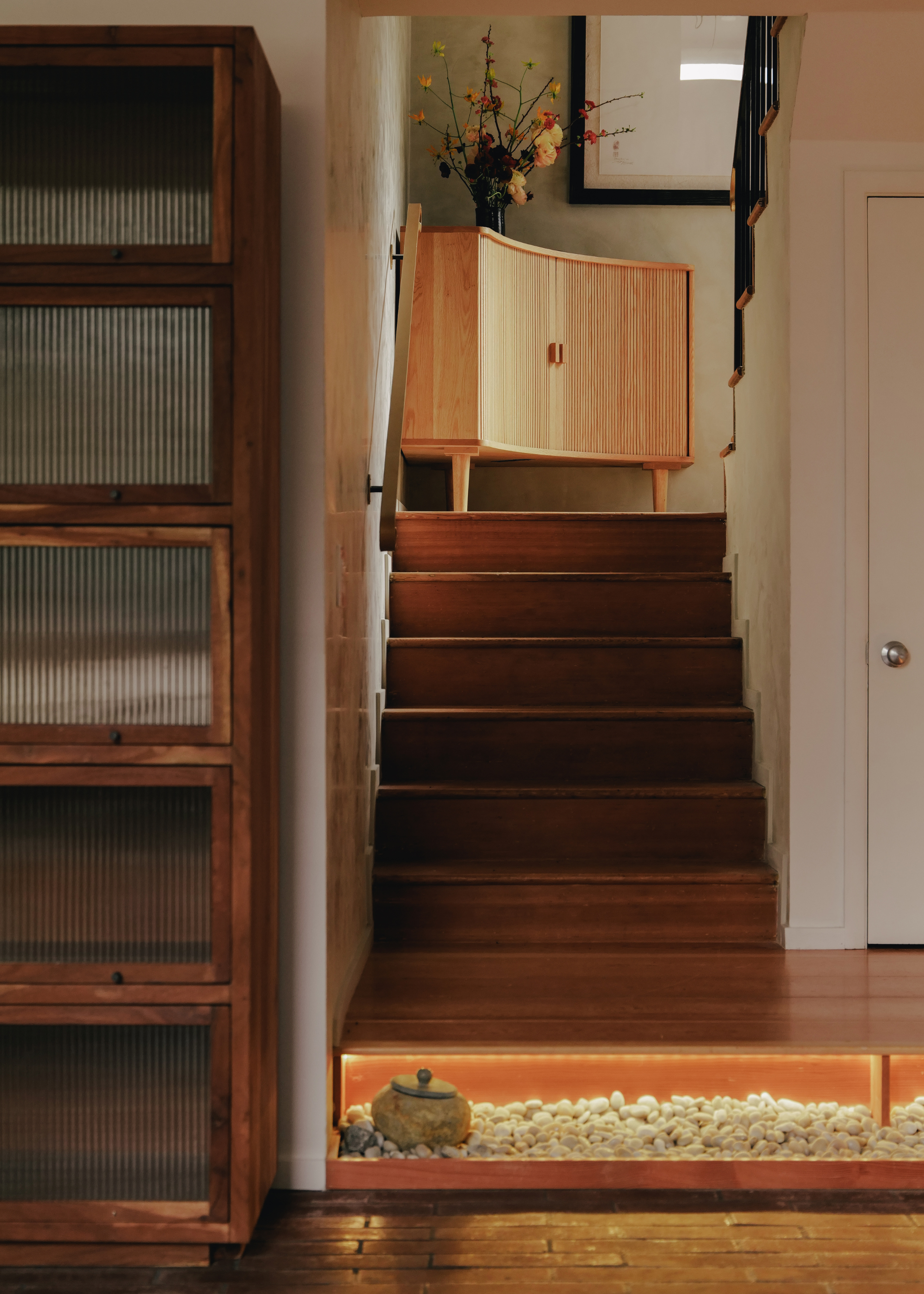An entryway with a wooden staircase, a fluted glass shoe cabinet, and a wooden ribbed sideboard with a vase of flowers