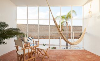 Interior view of the relaxing area in the Casa Mãe hotel. Large, airy space, with floor-to-ceiling windows looking over a palm tree, a sitting area with beach chairs in beige and blue, and a hammock.