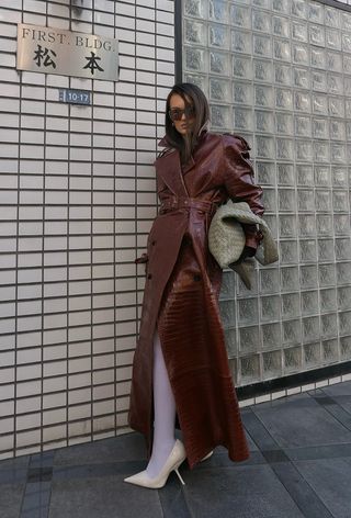 winter tight outfit shown in a photo of a woman wearing a long brown leather trench coat as a dress styled with white tights, white pumps, and an oversized sage green handbag