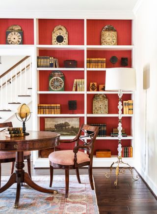 library with open shelves on red walls with dark wood floor and patterned rug with round pedestal table and dark wood carver chair and standard lamp with stairs in background