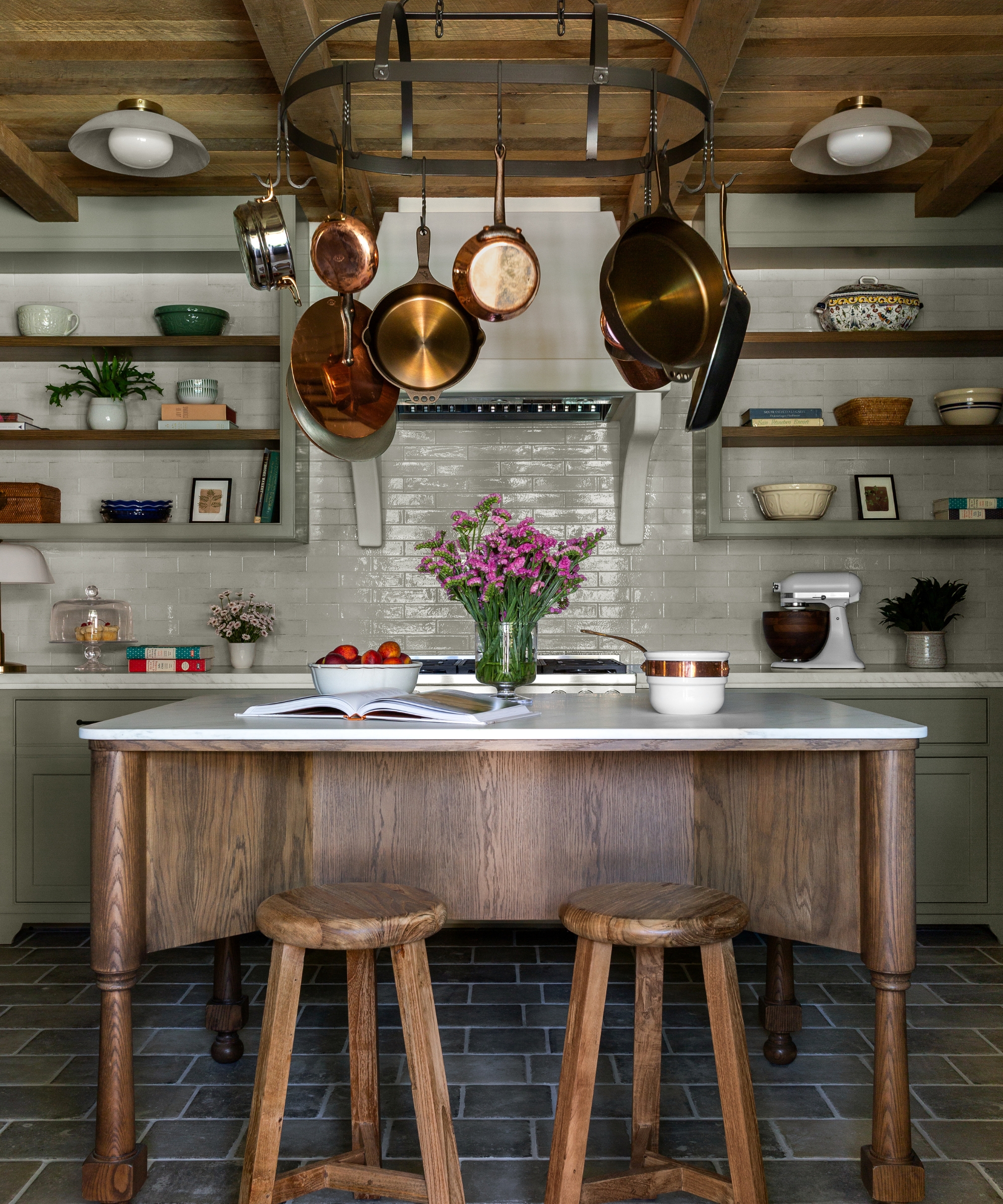 A baking kitchen with green tile walls, green cabinets, and a freestanding prep table-style wooden island. Above, an aged metal pot rack hangs from the ceiling, and wall shelves hold baking equipment