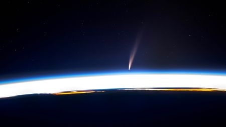 A photo taken from the ISS showing a bright comet and its streaking tail appearing to fall behind Earth's horizon