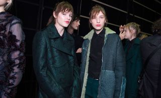 Four female models wearing looks from Costume National's collection. One model is wearing a semi-sheer piece with dark detail. Another model is wearing a dark green patterned coat. Next to her is a model wearing a black shirt, dark green trousers and a blue panelled coat with off-white coloured fur. And the fourth model is in the background wearing a dark green coat