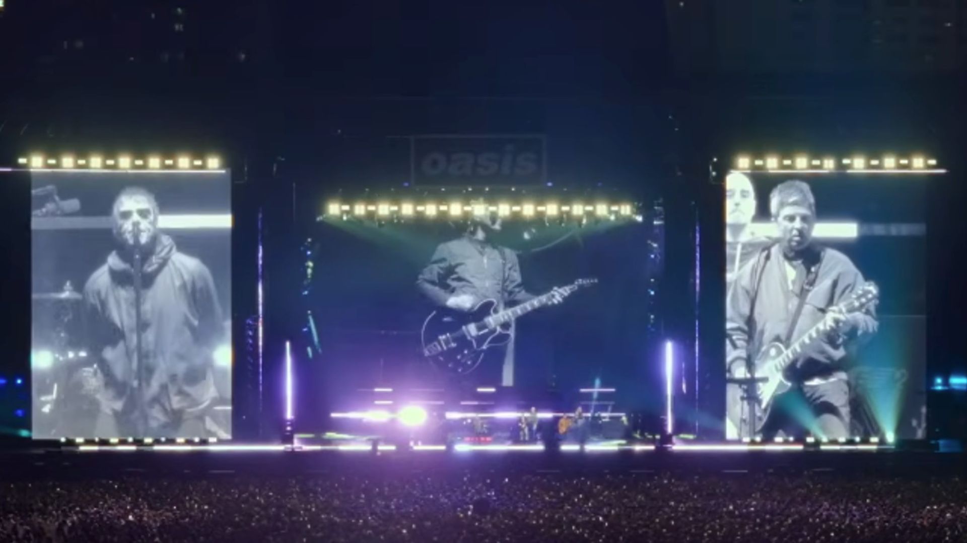 A wide shot of the Oasis concert stage with huge screens with the two Gallagher brothers on them in Oasis: Don't Look Back In Anger