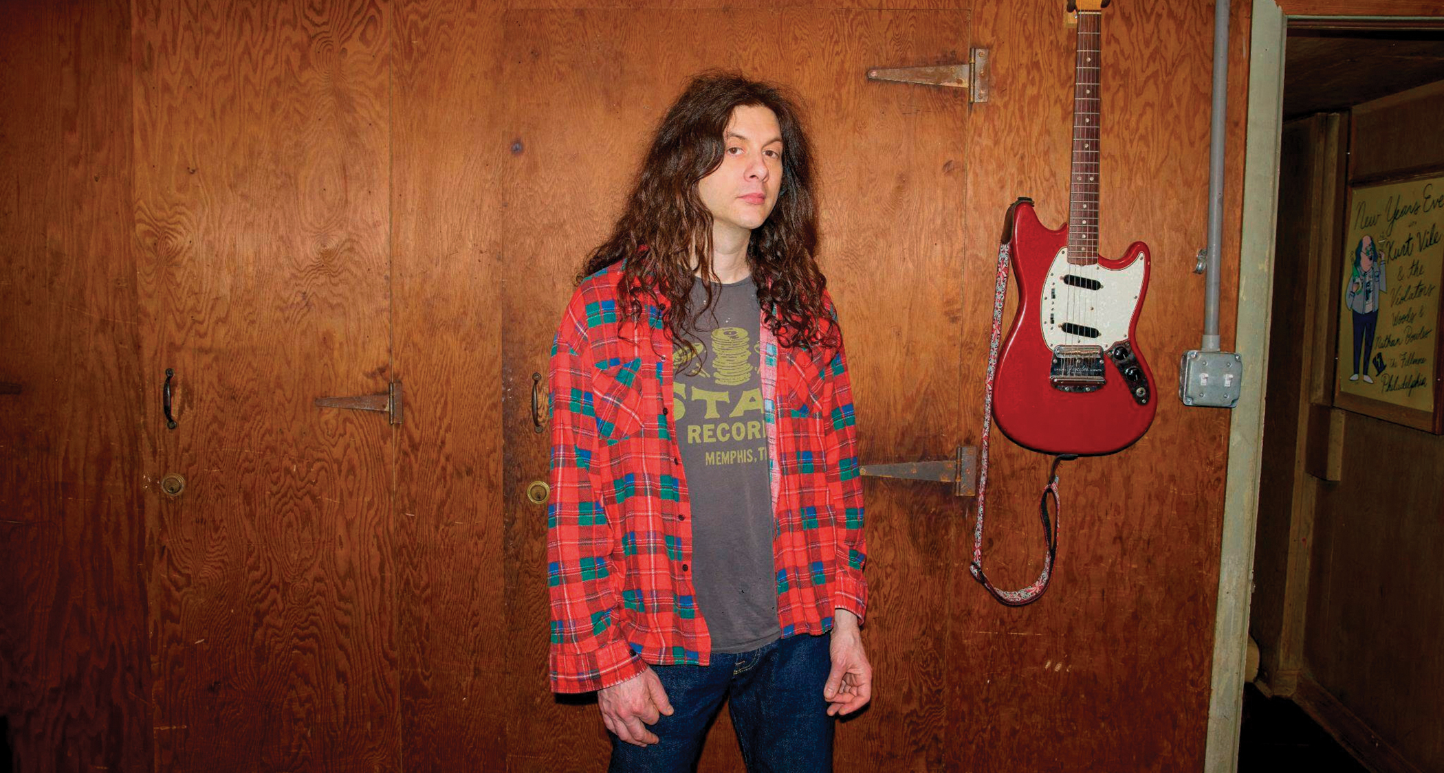 Kurt Vile pictured in front of a wall with a Fender Mustang