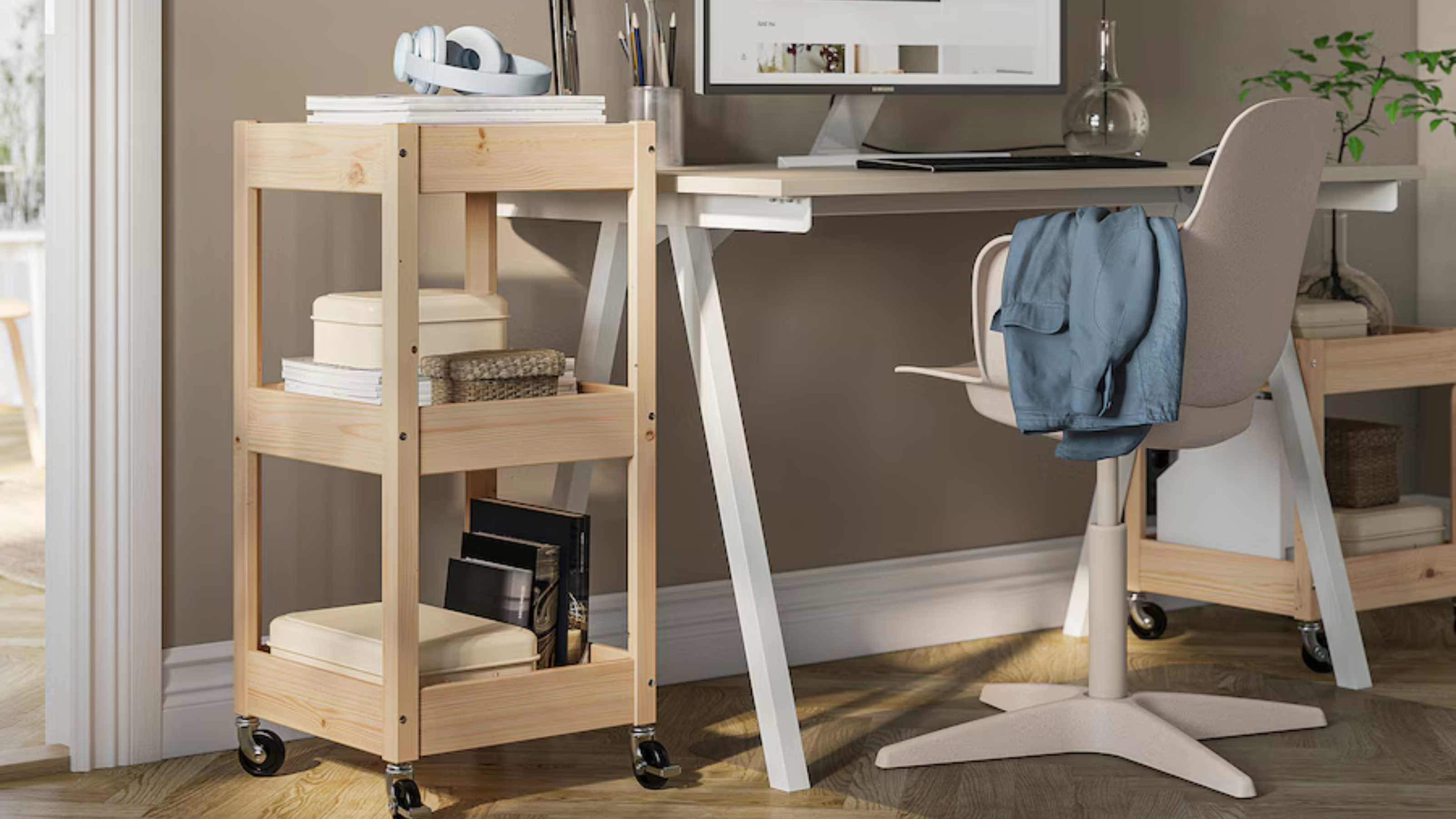 LOSHULT trolley next to a white desk in a home office. It matches the height of the desk and hold paperwork, stationery, books and headphones.