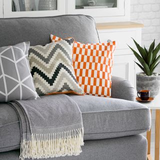 a white living room containing a grey sofa, with geometric pattern cushions in grey, white and orange and a grey throw