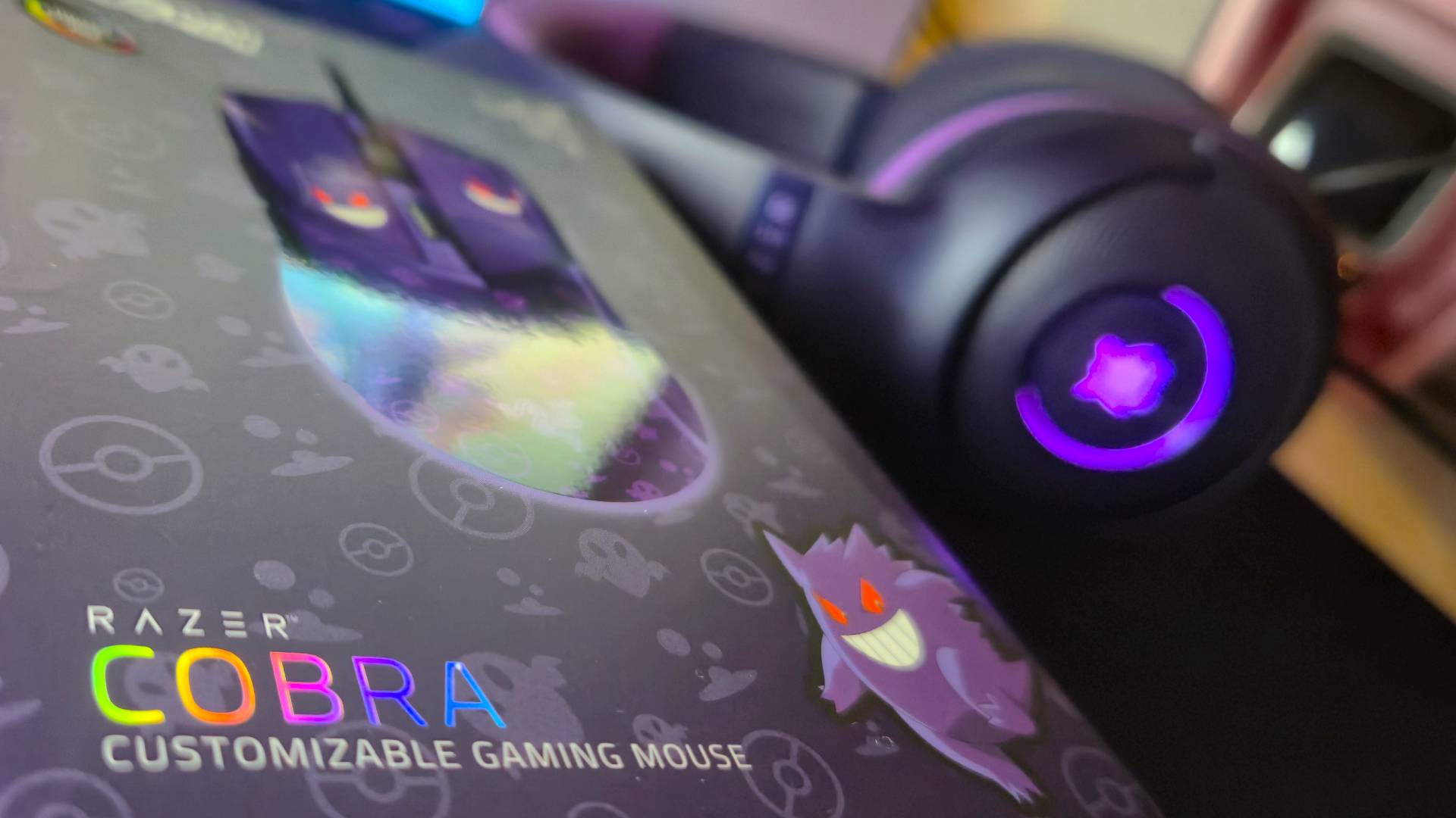Close up of the front of the box of the Razer Cobra Gengar gaming mouse, with a lit-up matching purple gaming headset in the background.