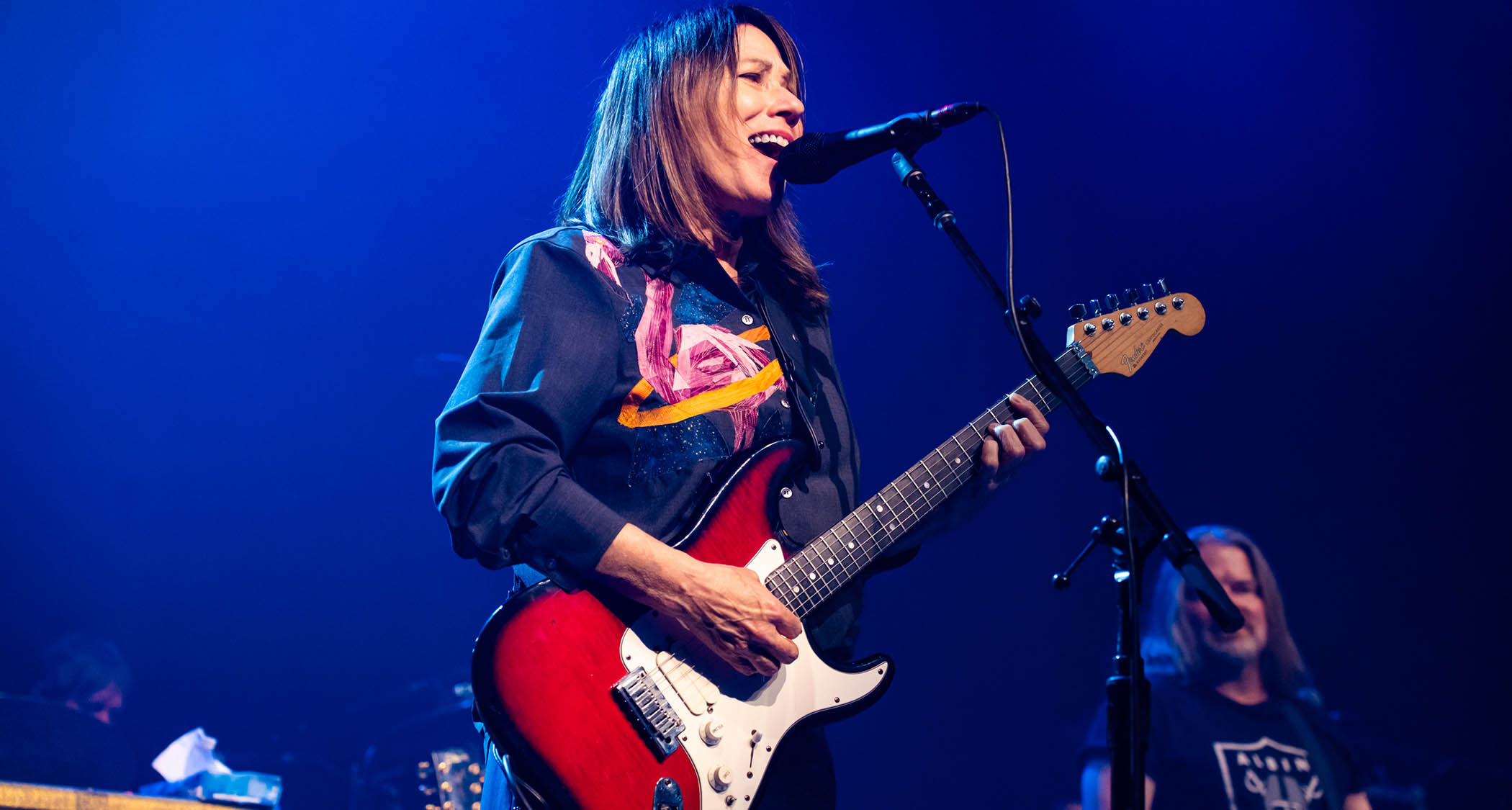 Kim Deal plays her Fender Strat as she performs at London&#039;s Roundhouse in June 2025, the stage lit up in blue.