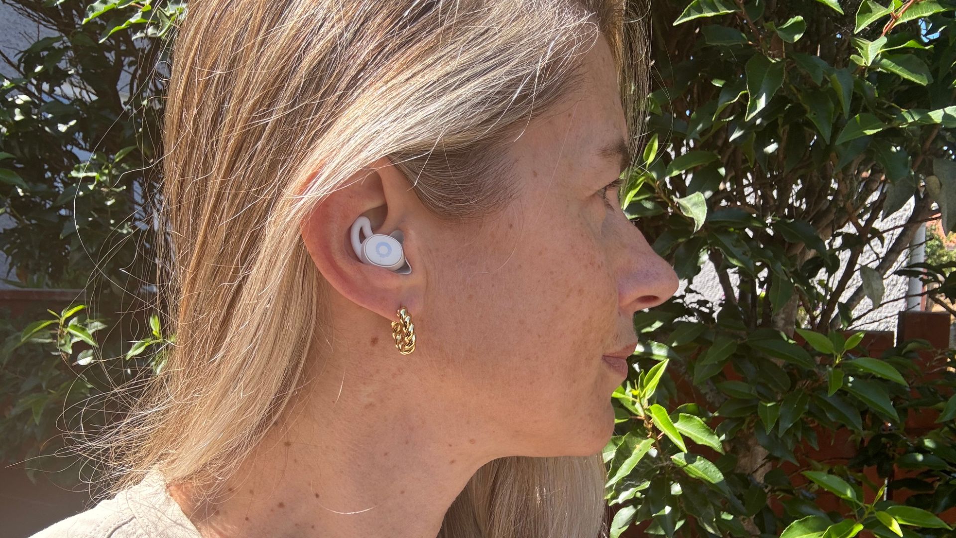 A blonde woman pictured from the neck up and standing in front of a green plant wears the white Ozlo Sleepbuds 2