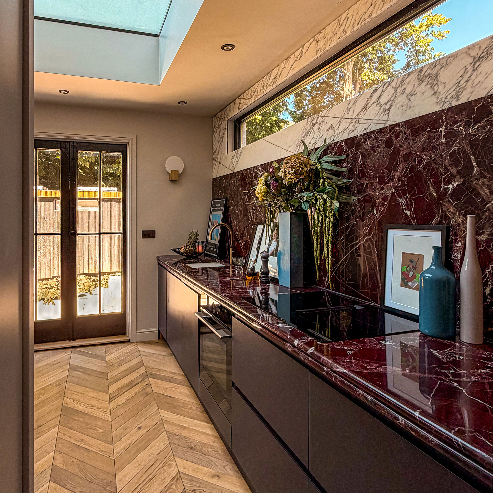 Kitchen with skylight, wooden chevron flooring, doors to garden, dark red cabinetry, red countertops and splashbacks topped with white marble