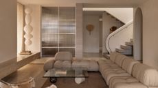 A beautiful modern minimalist living room with a long l-shaped couch, a glass coffee table, a paper lantern, curved archways, a curved staircase and a wall-mounted traditional fan