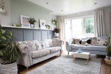 Living room with grey panelling to dado height, pastel green paint extending above and onto ceiling over shelving, pink sofa and grey sofa, wood flooring and grey rug