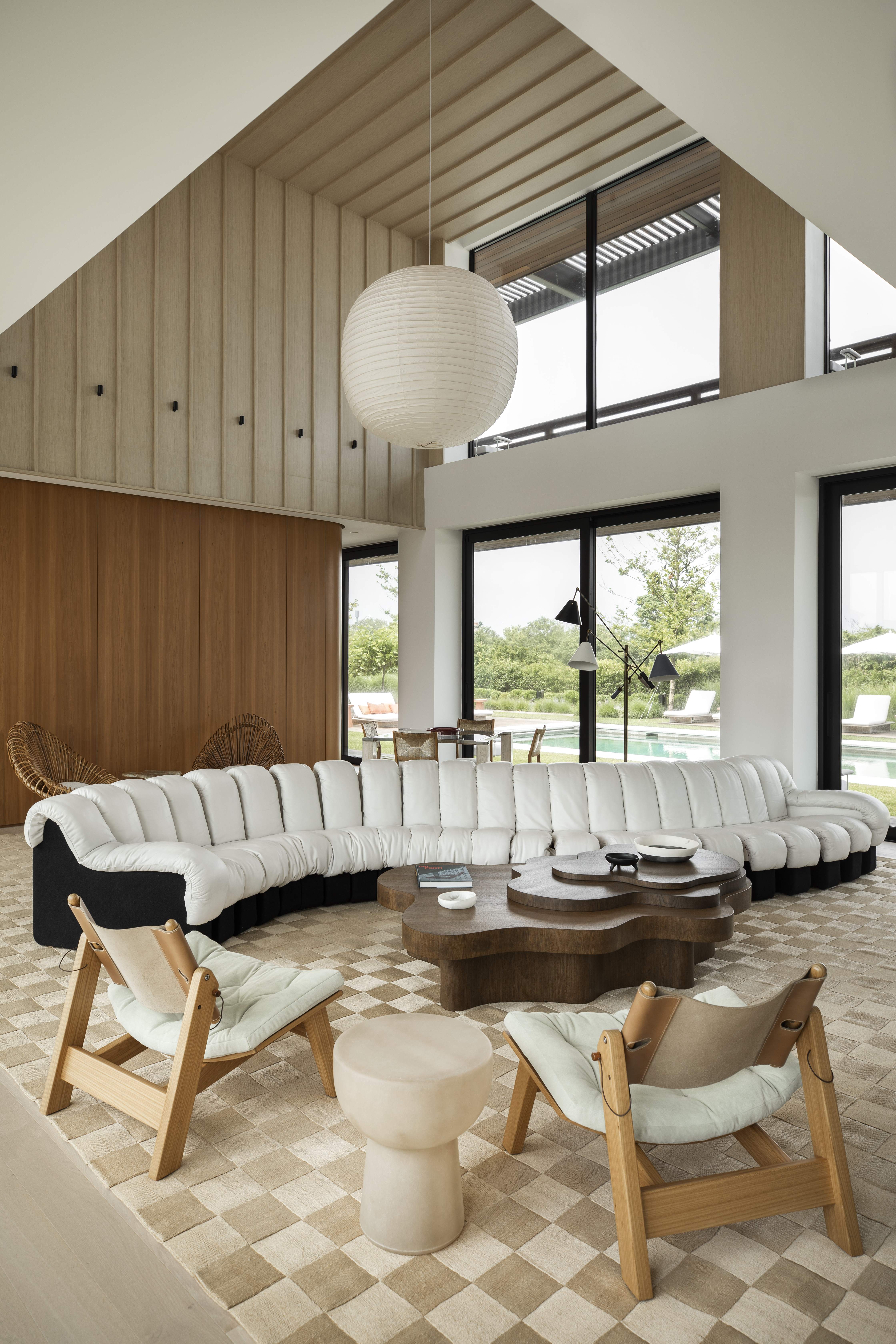 A high-ceilinged Japandi-style living room featuring a low-slung channel-tufted serpentine sofa, a wooden coffee table and chairs, along with an oversized lantern-style pendant light.