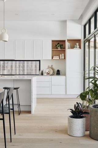 An all white kitchen with concrete accents, plants, metal frame windows and a kitchen island