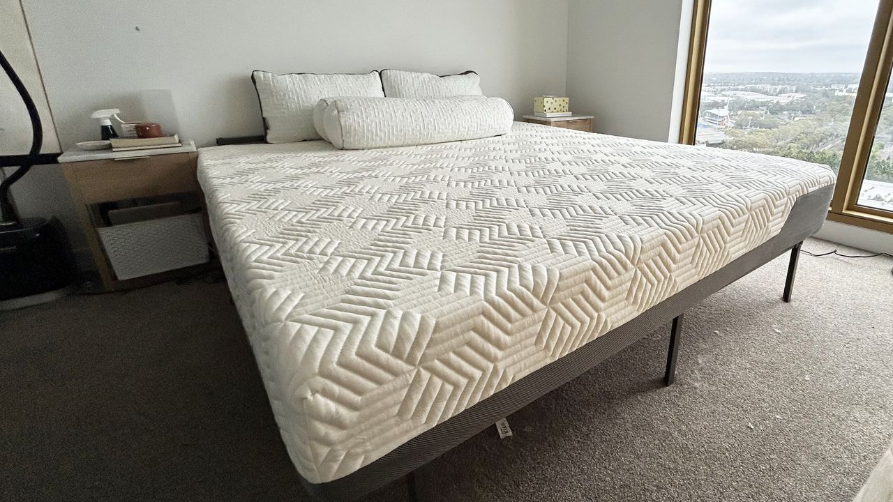 Valmori Hybrid II mattress on bed frame with four pillows in bedroom with two nightstands and window to the right
