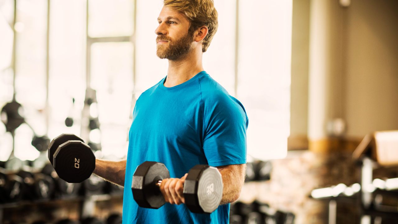 A man in a t-shirt performs biceps curls in a gym. He is standing, holding a pair of 20kg dumbbells in his hands; his elbows are bent and forearms parallel to the floor. 