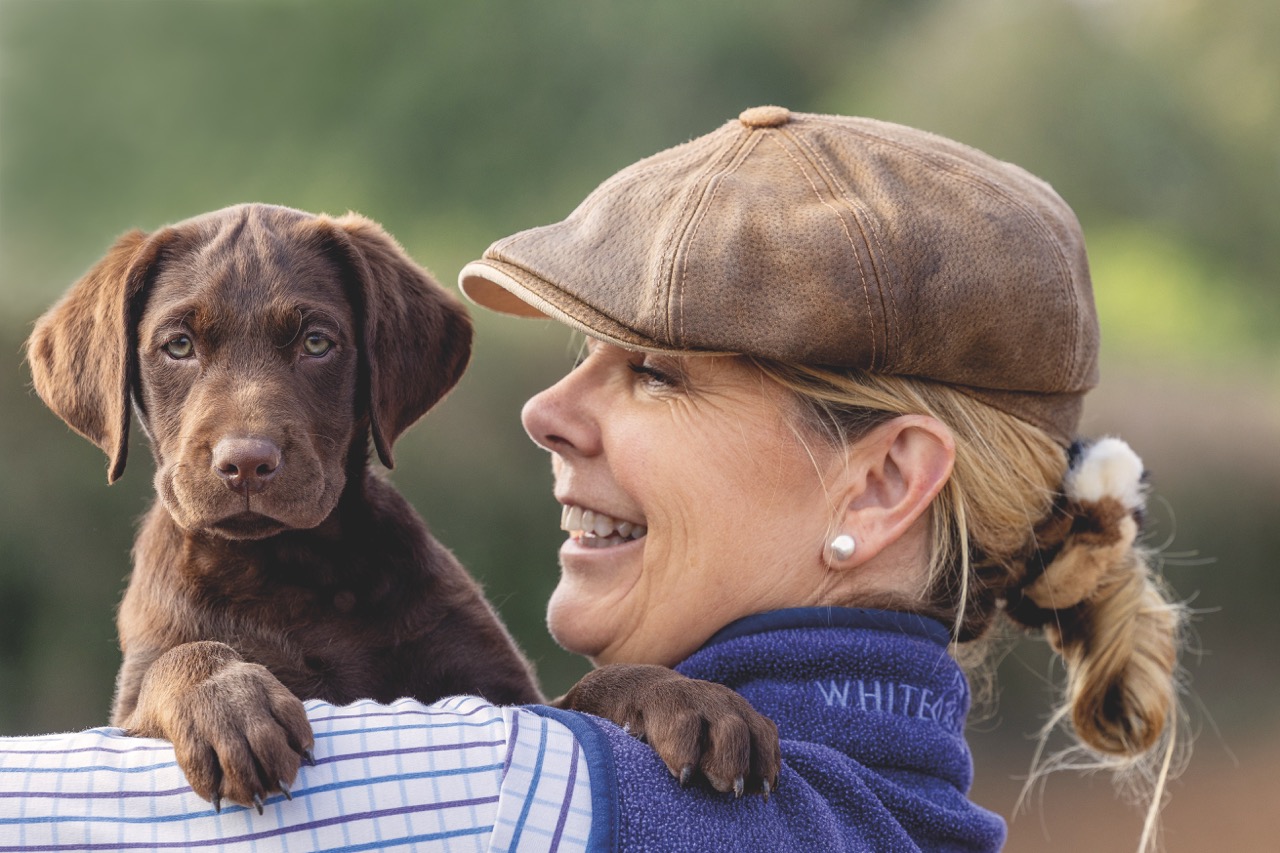 A woman smiles as she carries a young chocolate labrador puppy over her shoulder, the puppy resting its paws on her arm and looking directly at the camera.