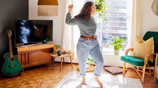 Woman dancing at home in living room to music in front of television, next to armchair and guitar, representing one way to do 10,000 at home