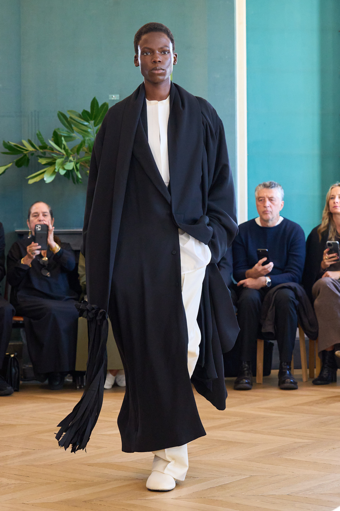 A model walking in the S/S 25 Carven show during Paris Fashion Week wearing a floor-length black coat with white pants and round toe ivory shoes.