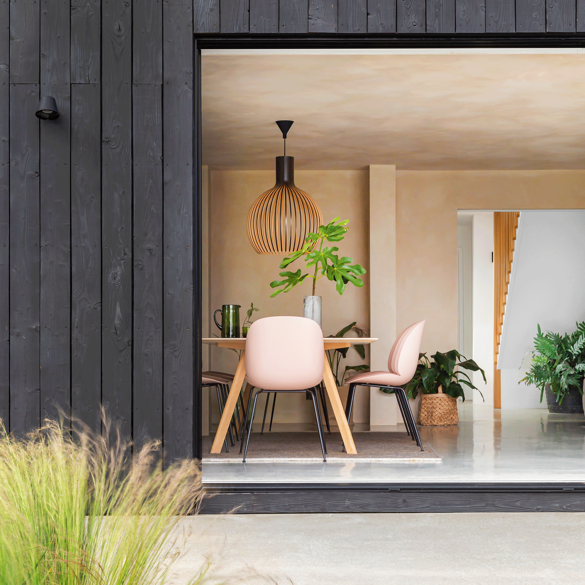 the exterior of a house covered in black timber cladding with a view into a dining area with pink dining chairs