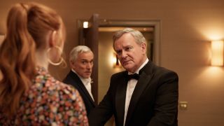 Karen Gillan in a brown floral dress as Madeline speaks to Hugh Bonneville in a dinner jacket as Douglas as Ben Miles in a dark suit as Toby stands between them in Douglas is Cancelle