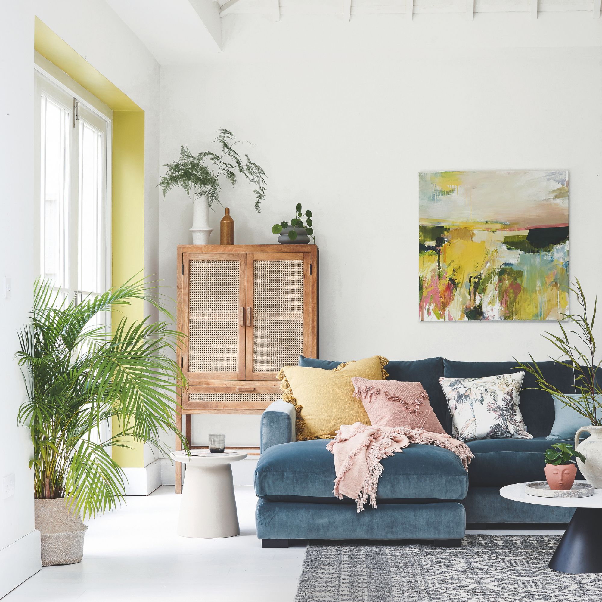 Light and airy living room, with a colourful print on the wall, a teal velvet sofa with pink and ochre coloured cushions, pink throw, wood and rattan cabinet and a plant on the floor by the window