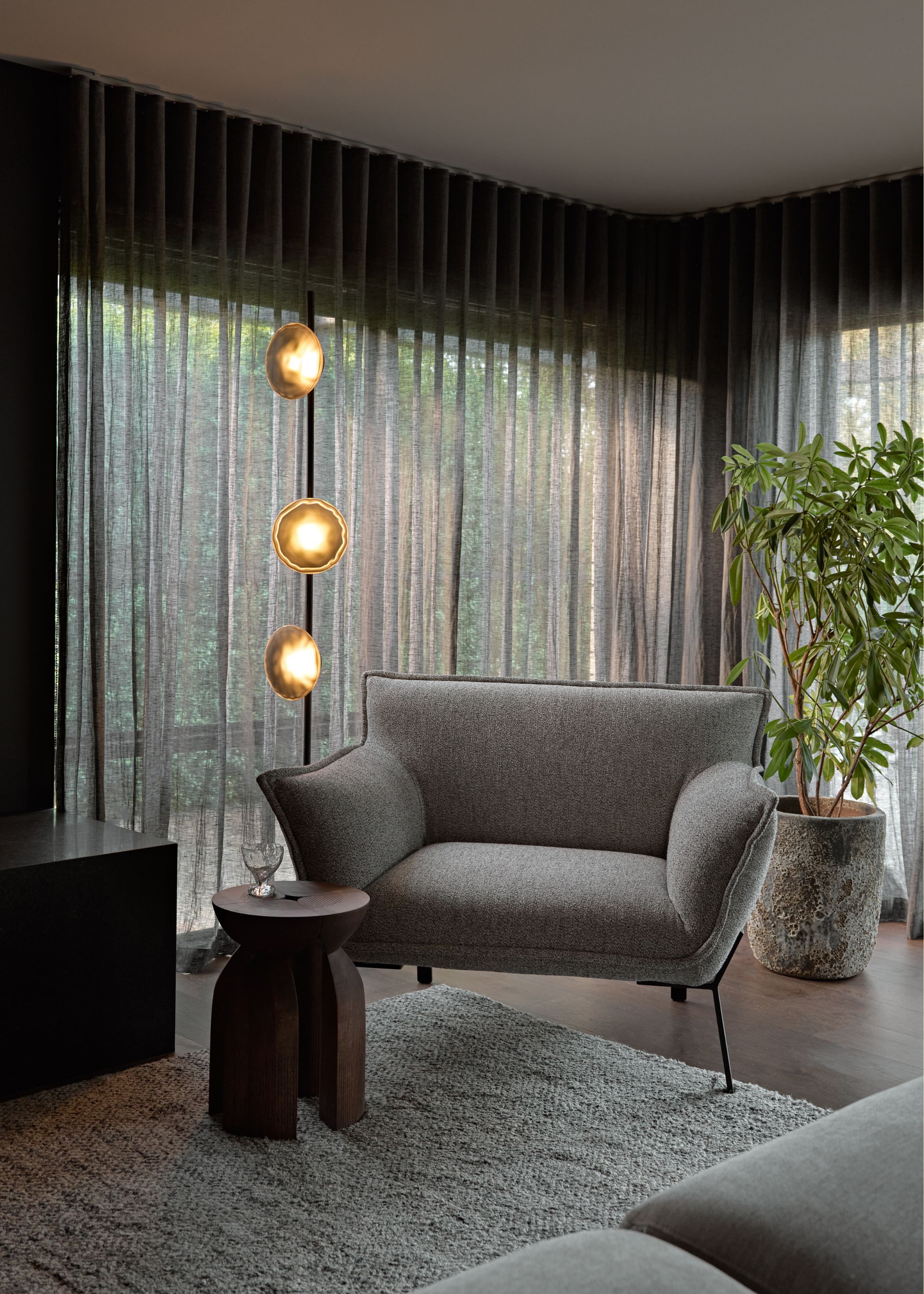 A muted living room with a multi-sconce floor lamp, a gray armchair, a wood side table, a potted tree, and a textured rug