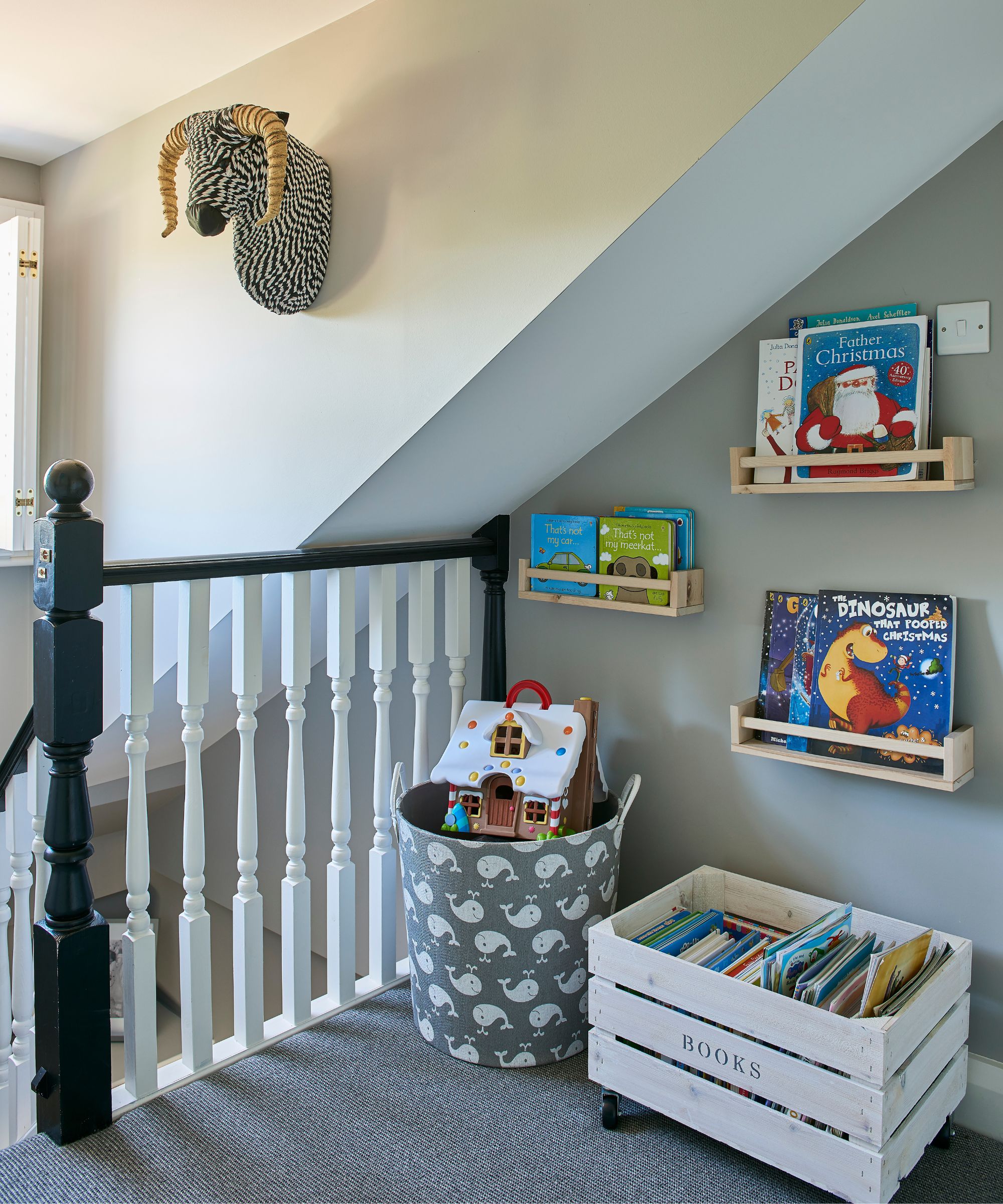 A neat and tidy space at the top of a black staircase with white banisters with organized kids books on shelves, and in a white wooden crate on the floor alongside a storage basket filled with floors and printed with whales. The floor is dark grey carpet, and the walls are pale grey.