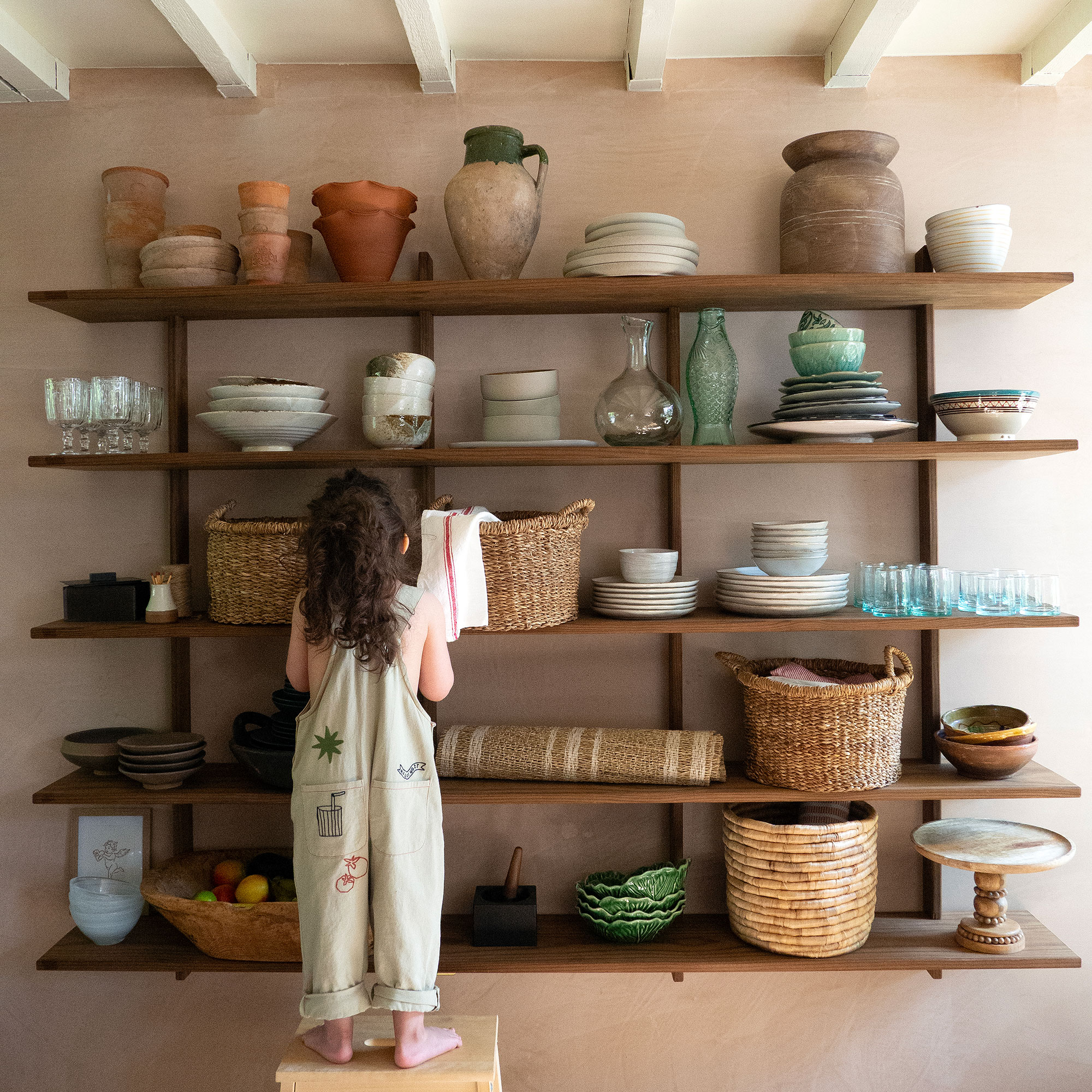 Small girl standing on stool in front of open wooden shelving filled with ceramics, glassware, jugs and baskets