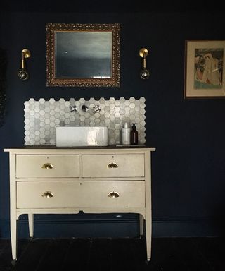 black bathroom ideas, black bathroom with black walls, cream vintage vanity unit with sink, pearlescent hexagonal tiled backsplash, ornate mirror and a pair of wall lights, artwork