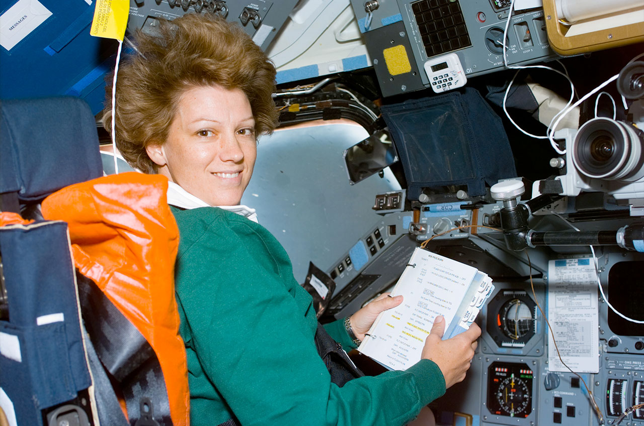 a woman in a green sweater reads a manual aboard the space shuttle