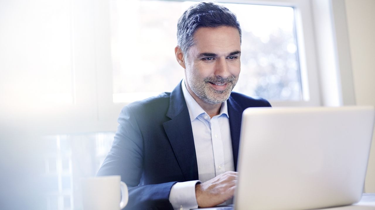 A businessman smiles as he works on his laptop at his desk in an office.