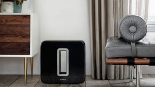 Sonos Sub i sort placeret på et gulv mellem en sofa og en kommode