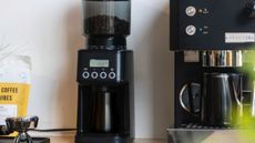 Barista & Co Core All Grind Plus Grinder on a countertop next to a coffee maker with a coffee bag to the left