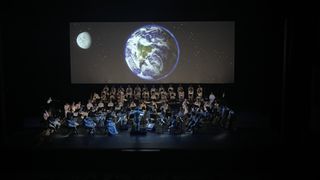 The orchestra and choir are lit with the Earth projected behind them.