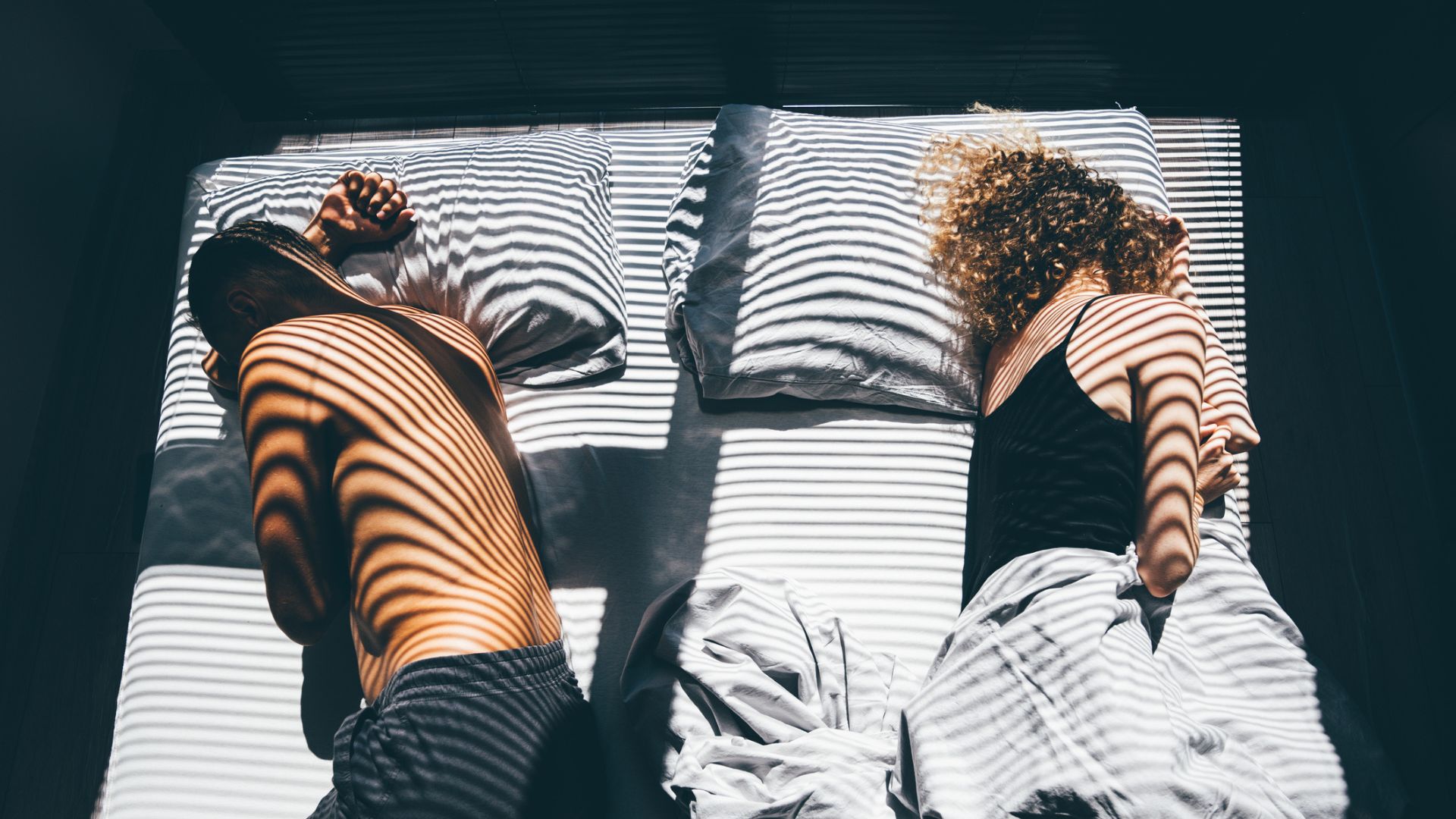 A couple sleeping on opposite sides of the bed as the sun streams through the blinds