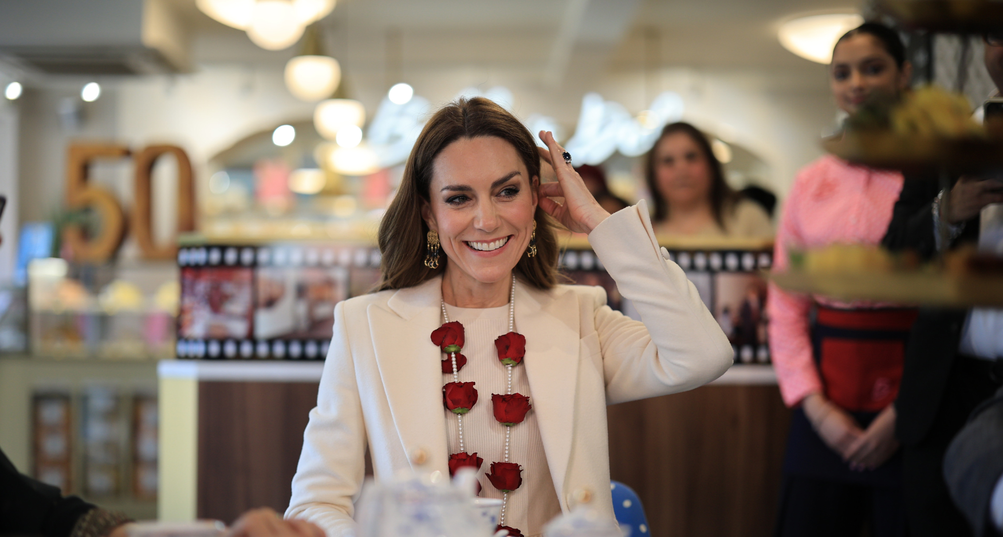 Princess Kate wearing a white coat and red rose garland smiling