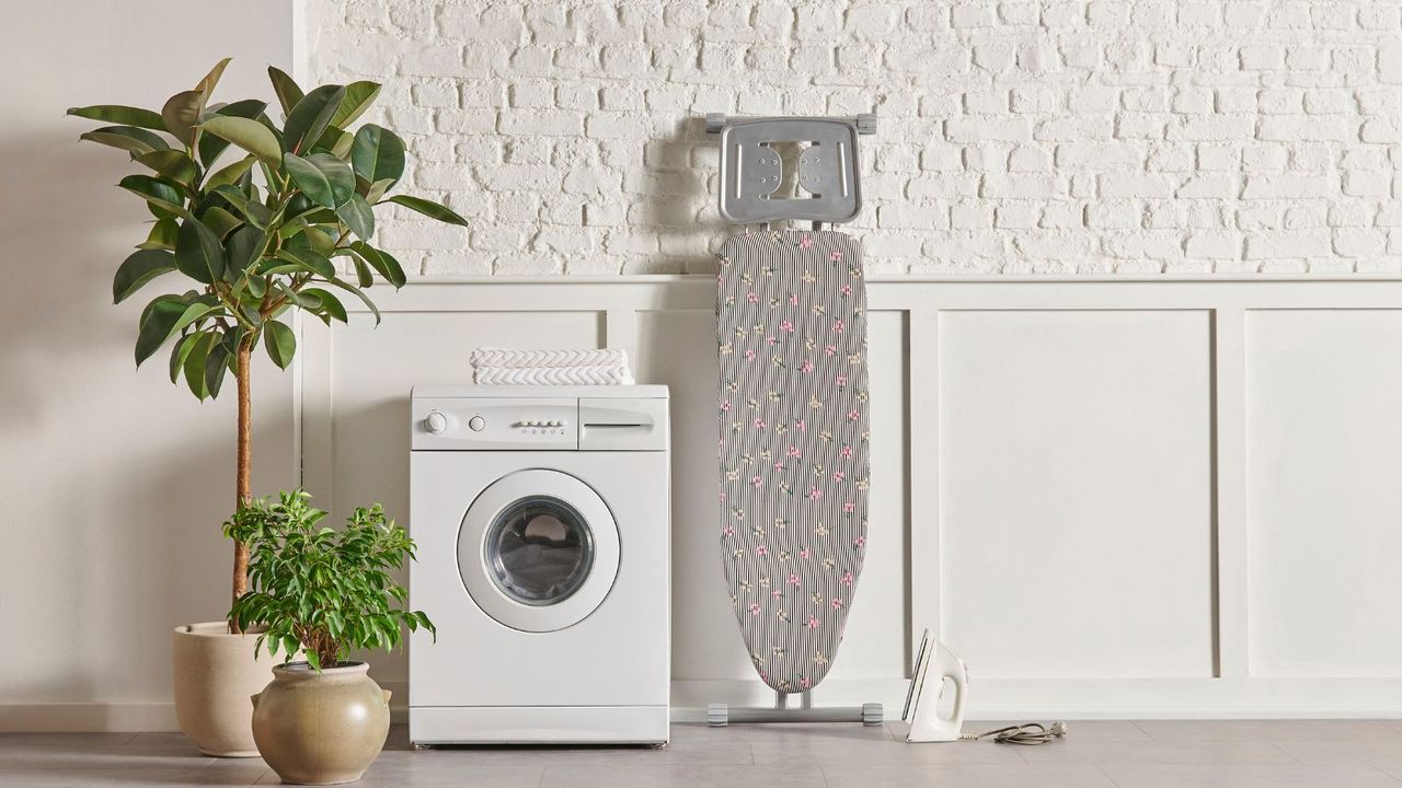 A white washer in the center of a room with a tree, plant pot on the left, ironing board to the right and cream paneling meets painted brick wall behind