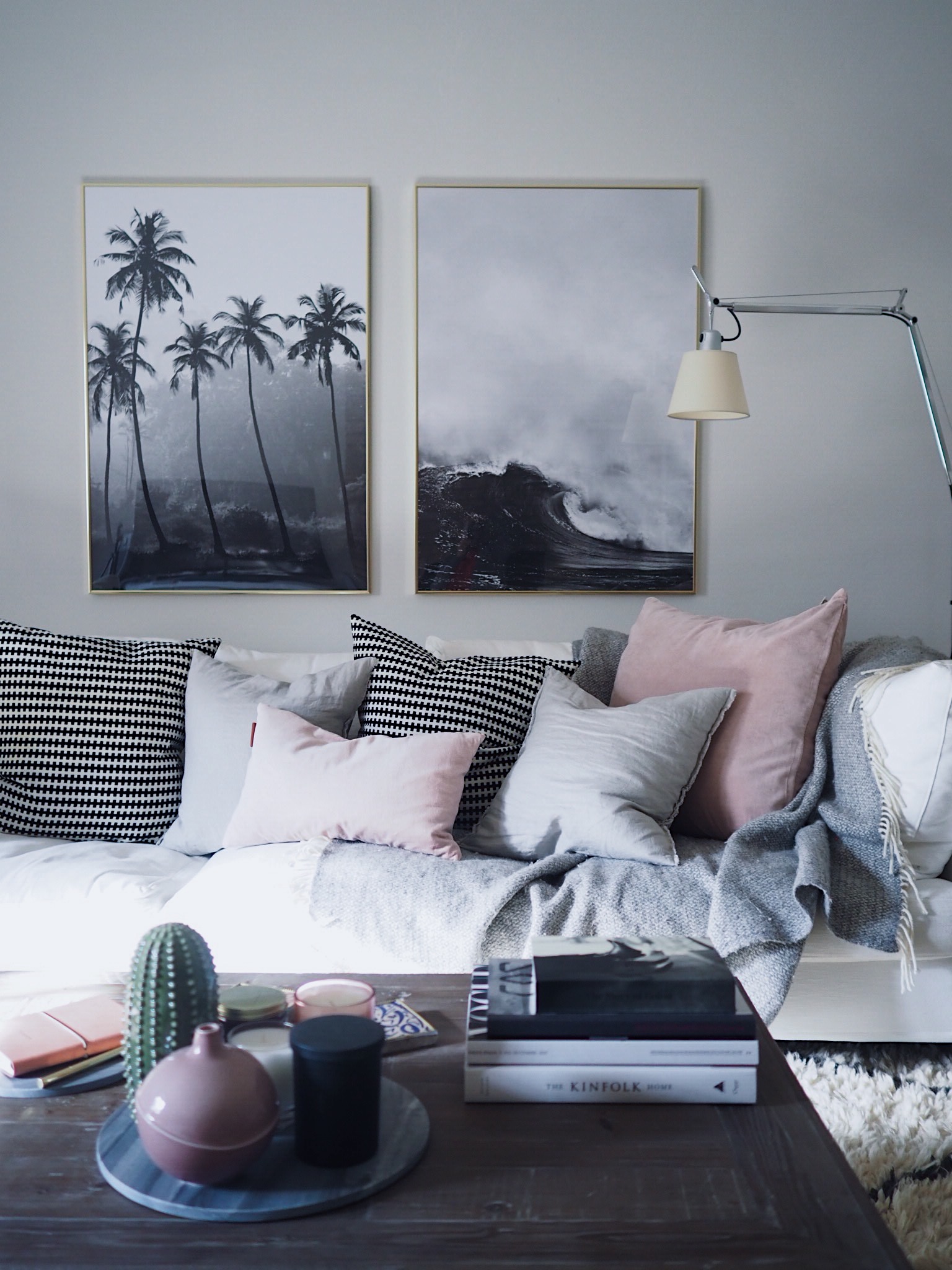 A grey living room with blush pink and striped monochrome cushions on sofa, palm-themed framed wall art and coffee table with decorative cacti ceramicware, and white floor lamp