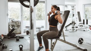 Woman performs dumbbell shoulder press sat on weights bench in gym