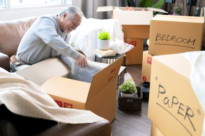 Horizontal medium shot of adult elder senior asian man male white beard sitting on floor preparing for moving to new house packing things into boxes and taping them in living room,home moving ideas concept