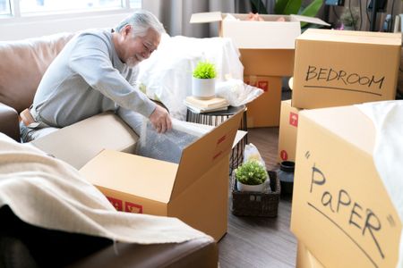 Horizontal medium shot of adult elder senior asian man male white beard sitting on floor preparing for moving to new house packing things into boxes and taping them in living room,home moving ideas concept